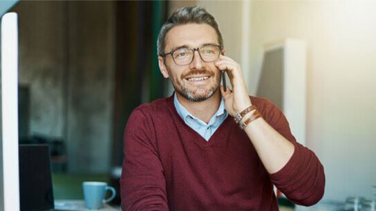 Lächelnder Mann mit Brille spricht am Telefon, sitzt an einem Schreibtisch mit Computer und Tasse.