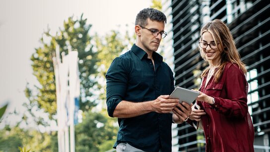 Ein Mann und eine Frau stehen im Freien und schauen gemeinsam auf ein Tablet, umgeben von Pflanzen und modernen Gebäuden.