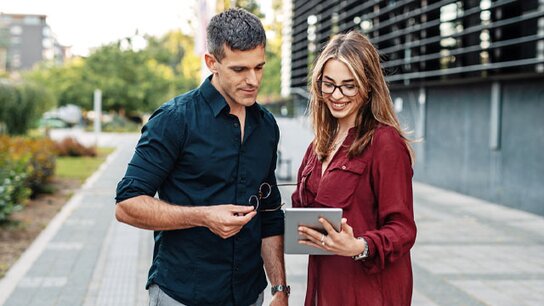Zwei Personen stehen im Freien und schauen gemeinsam auf ein Tablet, der Mann hält eine Sonnenbrille in der Hand.