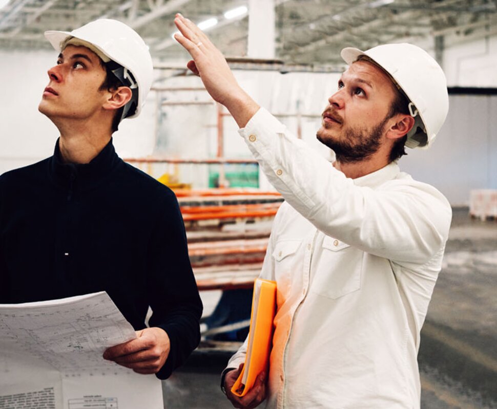 Zwei Bauarbeiter in Schutzhelmen betrachten Baupläne und diskutieren in einem großen, leeren Industriegebäude.