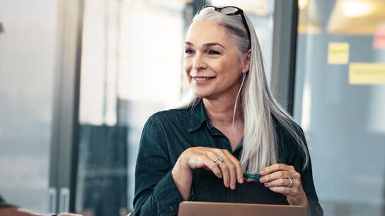 Ältere Frau mit langen grauen Haaren, lächelt und hält einen Stift, während ein Mann im Hintergrund ihr zusieht.