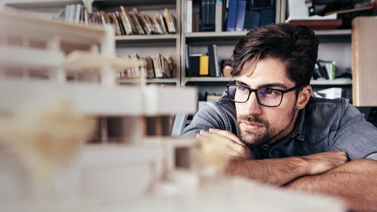 Junger Architekt betrachtet konzentriert ein Modell eines Gebäudes in einem Büro mit Regalen im Hintergrund.