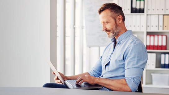 Mann mit kurzem Bart, sitzt an einem Tisch und arbeitet konzentriert am Laptop in einem modernen Büro.