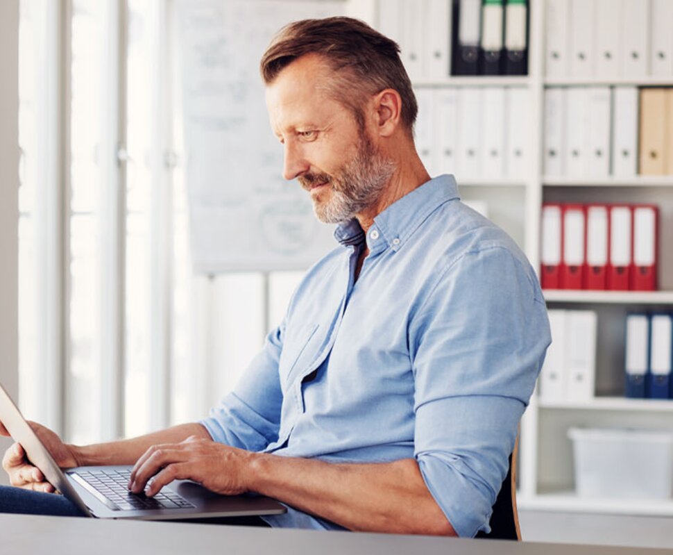 Mann mit kurzem Bart, sitzt an einem Tisch und arbeitet konzentriert am Laptop in einem modernen Büro.