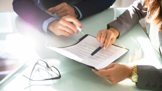 Zwei Personen diskutieren über einen Vertrag, während eine Hand auf einem Dokument zeigt und die andere einen Stift hält.