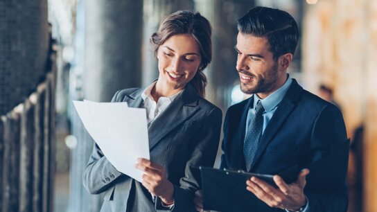 Ein Mann und eine Frau in Anzügen diskutieren lächelnd über Dokumente in einem modernen Büro.