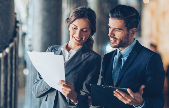 Ein Mann und eine Frau in Anzügen diskutieren lächelnd über Dokumente in einem modernen Büro.