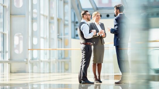 Drei Personen im Gespräch in einem modernen Bürogebäude mit großen Fenstern und hellem Licht.