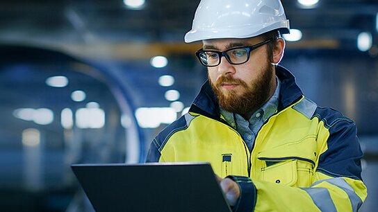 Ein Mann in Sicherheitskleidung und Helm arbeitet konzentriert an einem Laptop in einer industriellen Umgebung.