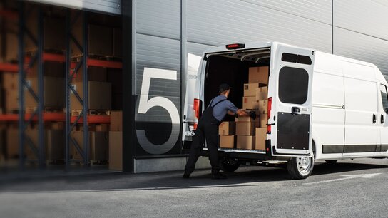 Lieferwagen wird entladen, während ein Arbeiter Kartons aus dem Rückraum herausnimmt, vor einem Lagerhaus mit Regalen.