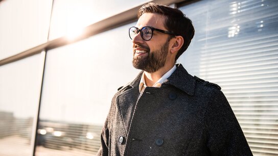 Lächelnder Mann in einer modernen Umgebung, trägt einen grauen Mantel und Brille, mit Sonnenlicht im Hintergrund.