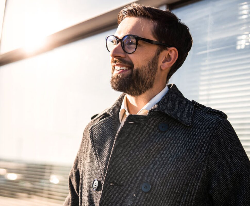 Lächelnder Mann in einer modernen Umgebung, trägt einen grauen Mantel und Brille, mit Sonnenlicht im Hintergrund.