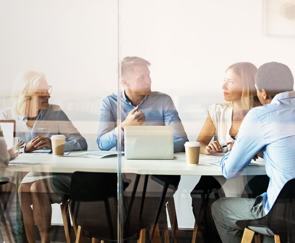 Sechs Personen in einem Konferenzraum, die an einem Tisch sitzen und ein Gespräch führen, mit Laptops und Kaffeetassen.