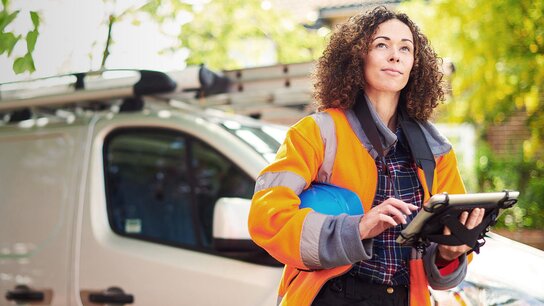 Frau in Sicherheitskleidung mit Helm, die auf einem Tablet arbeitet, steht neben einem Lieferwagen in einer grünen Umgebung.