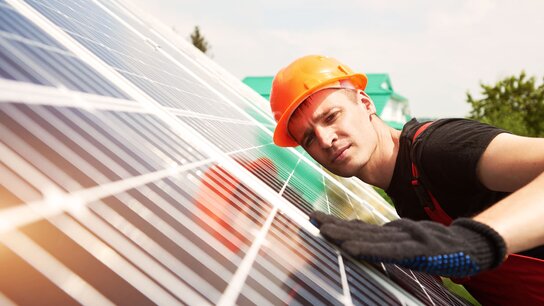 Ein Arbeiter in orangefarbener Schutzhelm und Handschuhen überprüft eine Solaranlage auf einem Dach.