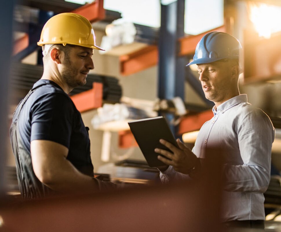 Zwei Männer in Schutzhelmen diskutieren in einem Lager, einer hält ein Tablet in der Hand.