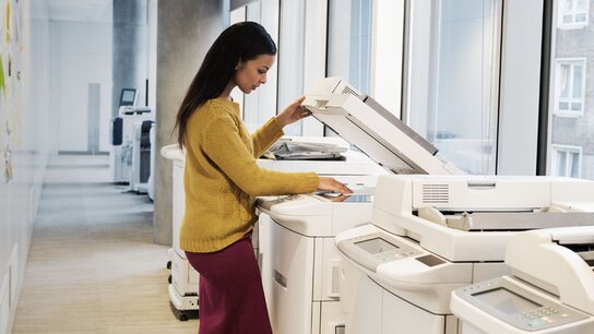 Frau in gelbem Pullover stehend an einem Kopierer in einem modernen Büroraum mit großen Fenstern.