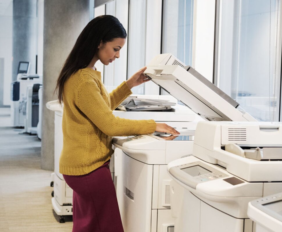 Frau in gelbem Pullover stehend an einem Kopierer in einem modernen Büroraum mit großen Fenstern.