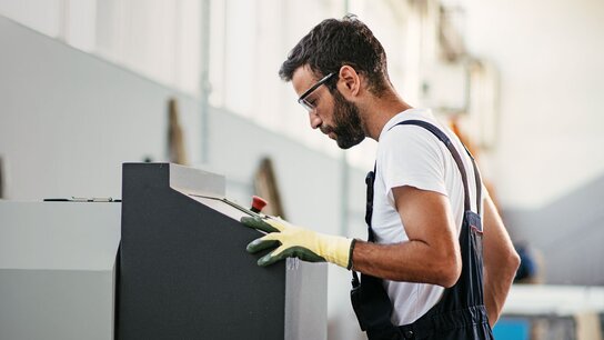 Ein Mann in Arbeitskleidung und Schutzbrille konzentriert sich auf eine Maschine in einer Industrieumgebung.