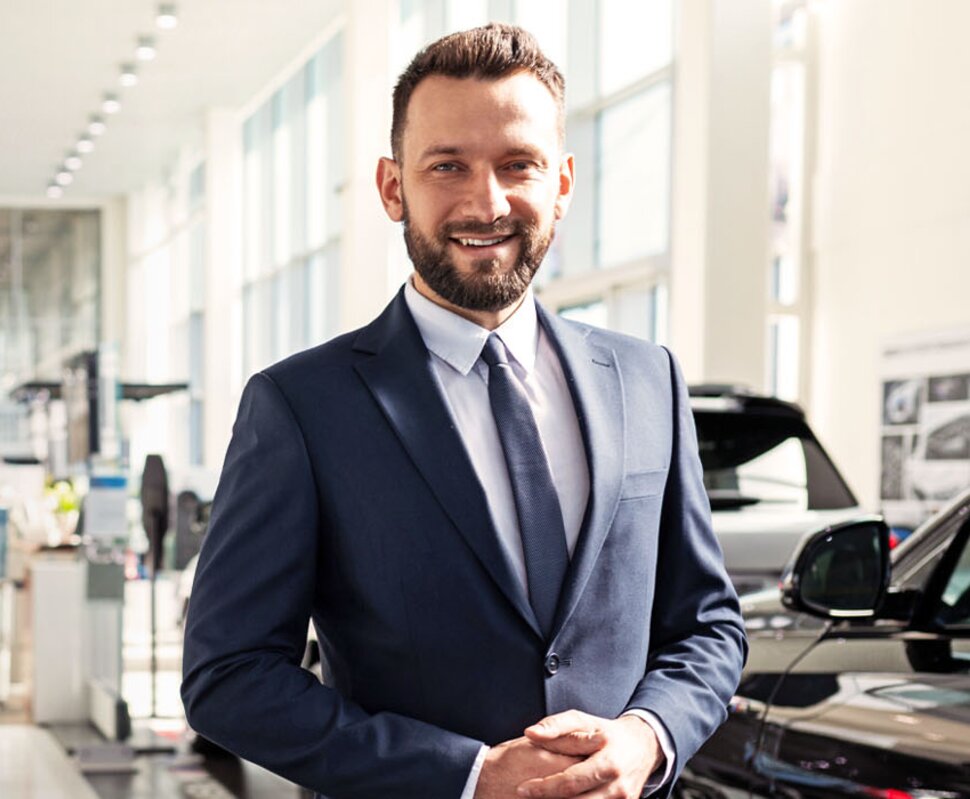 Mann im Anzug steht in einem Autohaus, umgeben von mehreren Fahrzeugen, und lächelt freundlich.