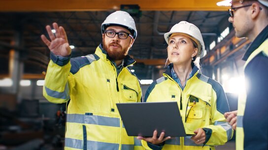 Drei Bauarbeiter in Helmen und reflektierender Kleidung besprechen Pläne mit einem Laptop in einer Lagerhalle.