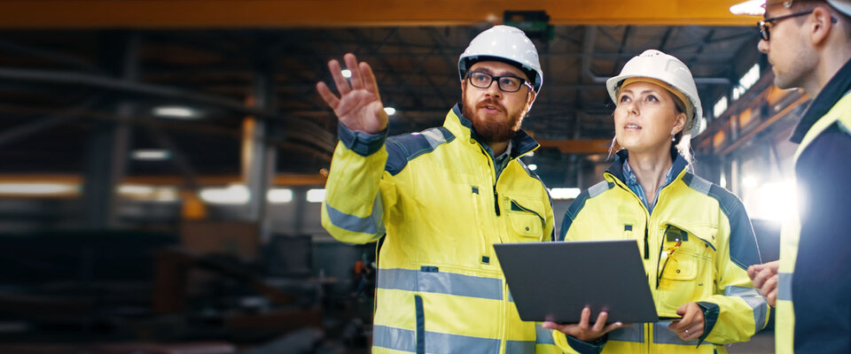 Drei Bauarbeiter in Helmen und reflektierender Kleidung besprechen Pläne mit einem Laptop in einer Lagerhalle.