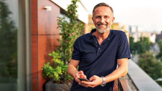 Lächelnder Mann im Polohemd steht auf einem Balkon, hält ein Smartphone in der Hand, im Hintergrund Pflanzen und Stadtblick.