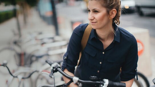 Junge Frau mit braunem Haar, trägt ein schwarzes Hemd und einen Rucksack, steht neben einem Fahrrad auf einer Stadtstraße.