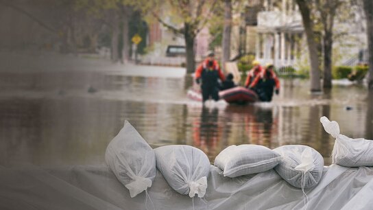 Sandsäcke liegen vor überflutetem Gebiet, während Rettungskräfte in einem Boot Hilfe leisten.