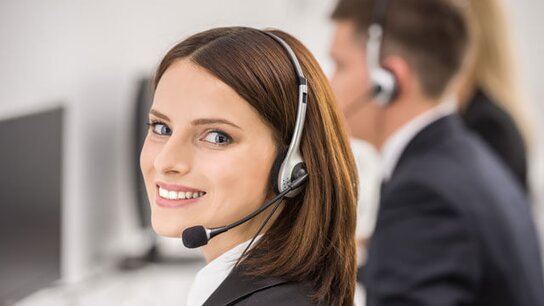 Frau mit Kopfhörern und Mikrofon lächelt zufrieden in einem Callcenter, im Hintergrund weitere Mitarbeiter.