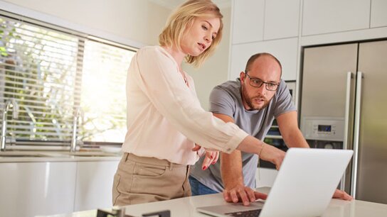 KI-generiertes Bild: Ein Mann und eine Frau stehen in einer modernen Küche und betrachten gemeinsam den Bildschirm eines Laptops.