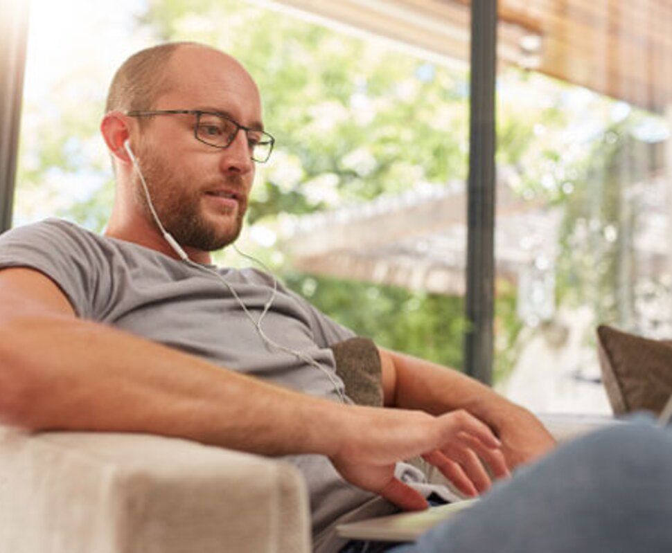 Mann mit Kopfhörern sitzt entspannt auf einem Sofa und arbeitet konzentriert am Laptop. Helles, freundliches Ambiente.