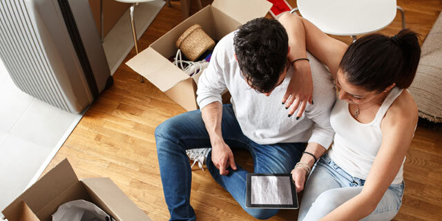 Zwei Personen sitzen auf dem Boden, umschlungen und betrachten gemeinsam ein E-Book auf einem Tablet, umgeben von Umzugskartons.