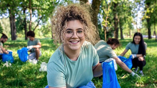 Junge Frau mit lockigem Haar lächelt und sammelt Müll im Park, während andere Helfer im Hintergrund aktiv sind.