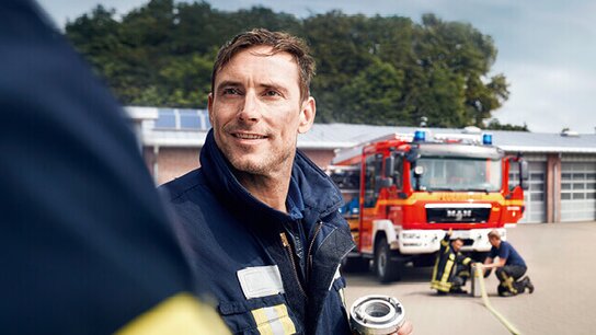 Lächelnder Feuerwehrmann in Uniform vor einem Feuerwehrauto, während Kollegen an einem Schläuch arbeiten.