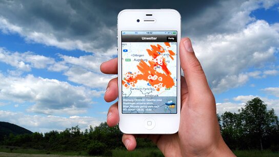 Hand hält ein Smartphone, auf dem eine Wetter-App mit einer Karte und Wolkenformationen angezeigt wird.