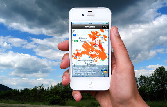 Hand hält ein Smartphone, auf dem eine Wetter-App mit einer Karte und Wolkenformationen angezeigt wird.
