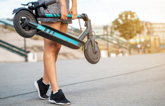 Junge Person trägt einen blauen E-Scooter in einer urbanen Umgebung auf einem gepflasterten Weg.