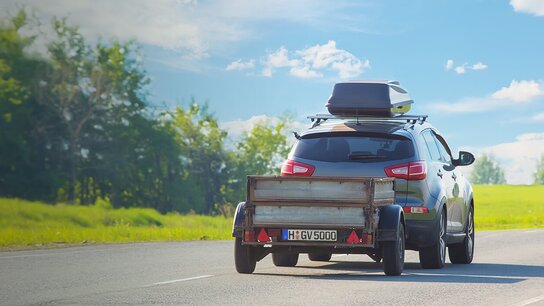 Ein SUV mit Anhänger auf einer Landstraße, umgeben von grünen Bäumen und klarem Himmel.