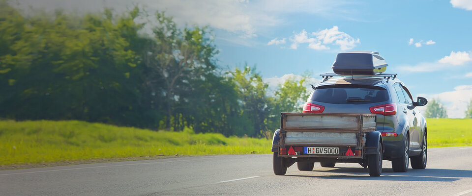 Ein SUV mit Anhänger auf einer Landstraße, umgeben von grünen Bäumen und klarem Himmel.