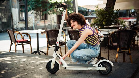 Eine Person mit lockigem Haar sitzt auf einem weißen Elektroroller in einem Straßencafé.