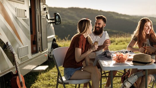 Vier Freunde sitzen an einem Tisch im Freien, genießen Snacks und Getränke, während ein Wohnmobil im Hintergrund steht.