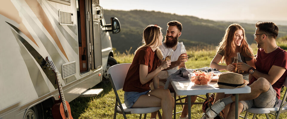 Vier Freunde sitzen an einem Tisch im Freien, genießen Snacks und Getränke, während ein Wohnmobil im Hintergrund steht.