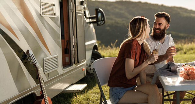 Paar sitzt im Freien vor einem Wohnmobil, genießt Snacks und Getränke, umgeben von Natur. Entspannte Stimmung.