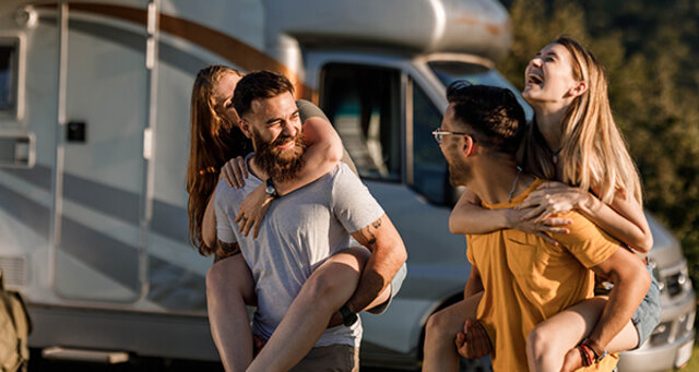 Vier Freunde lachen und haben Spaß, während zwei Frauen auf den Schultern ihrer männlichen Freunde vor einem Wohnmobil sitzen.