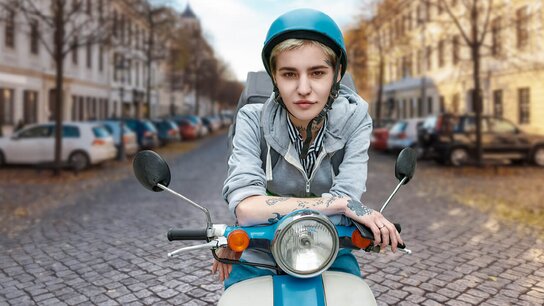 Junge Person auf einem Roller in einer urbanen Umgebung mit gepflasterter Straße und parkenden Autos im Hintergrund.