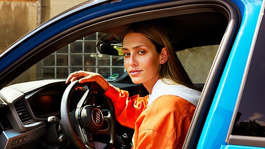 Lachender älterer Mann mit Brille sitzt am Steuer eines Autos und blickt freundlich nach außen. Sonnenlicht schimmert hinein.
