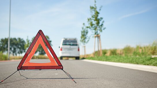 Warndreieck auf einer Straße, im Hintergrund ein parkendes Fahrzeug und grüne Bäume. Heller blauer Himmel.