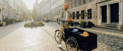 Mann mit Beanie und Rucksack steht neben einem Lastenrad in einer sonnigen, ruhigen Straßenansicht.