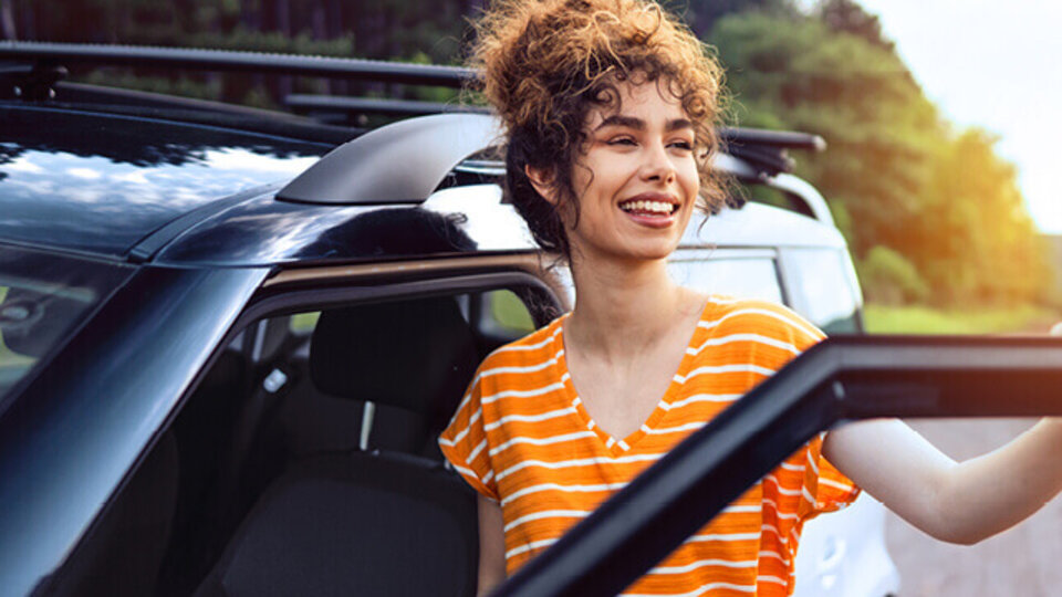 Lächelnde Frau mit lockigem Haar, die aus einem Auto steigt, im Hintergrund Bäume und ein weiteres Fahrzeug.